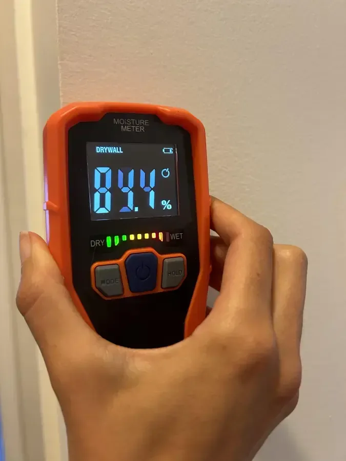 A handheld moisture meter pressed against an interior wall displays a reading