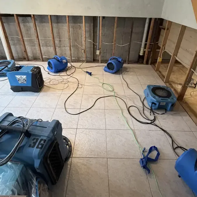 Blue air movers and a dehumidifier surround exposed wall framing above a tiled floor