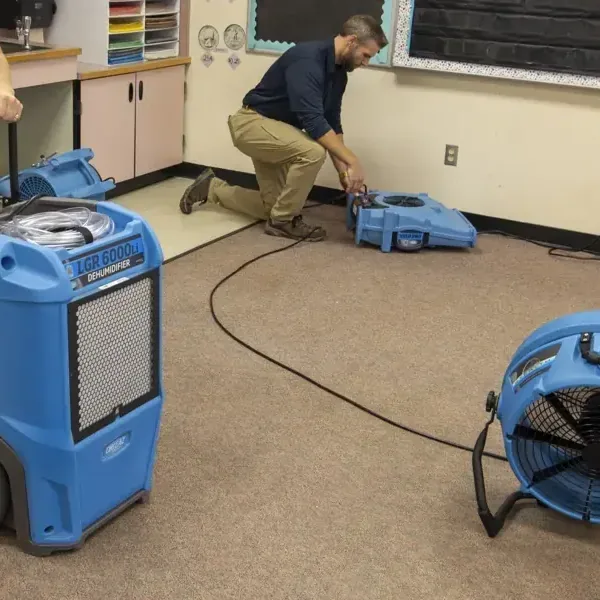 A technician adjusts a blue air mover beside a dehumidifier in a carpeted room