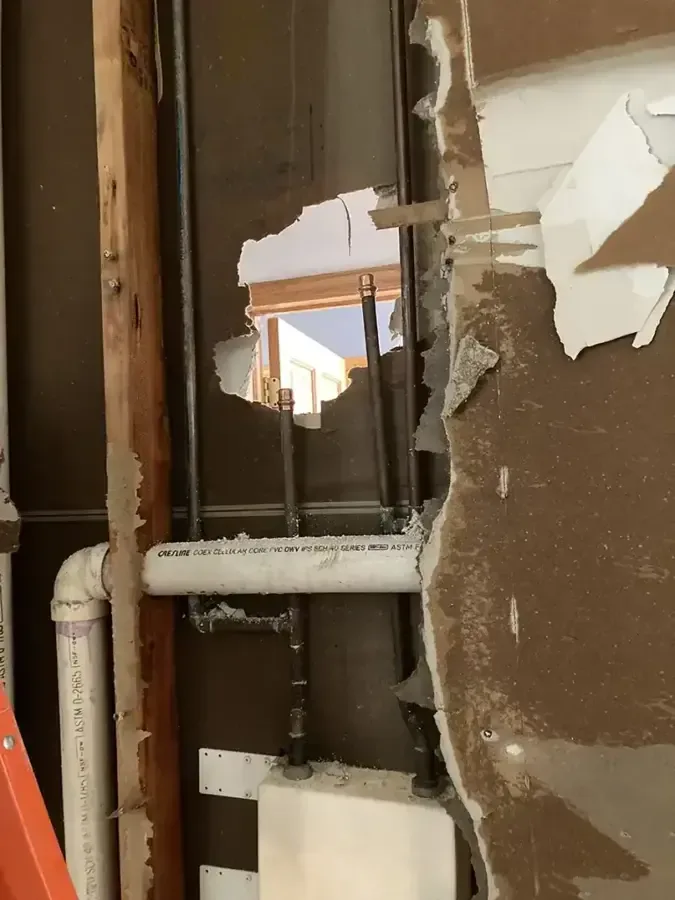 An opening in damaged wallboard exposes vertical framing and intersecting white plumbing pipes
