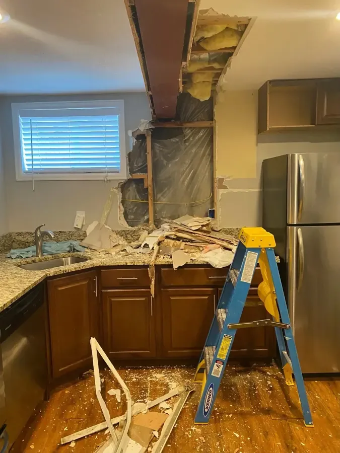 A ladder stands beside kitchen cabinets beneath a ceiling opened around exposed framing