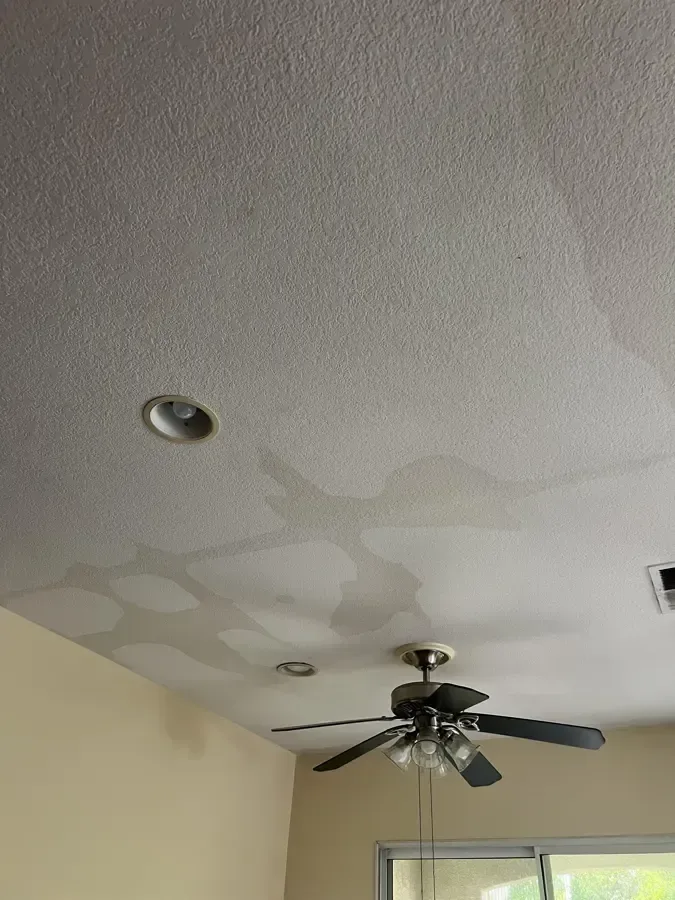 Water stains spread across a white ceiling above a ceiling fan and recessed lights