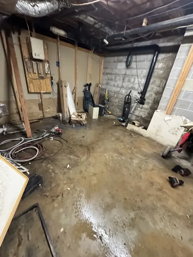 Water and debris cover a basement floor beside exposed drain pipes after a sewage backup