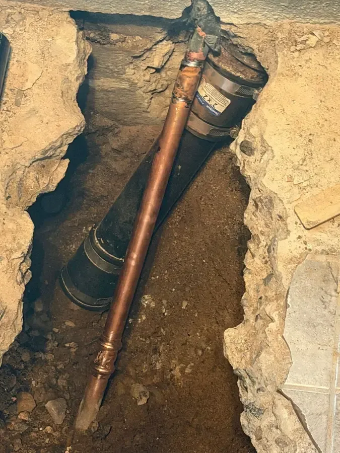 A copper pipe runs beside a drain pipe inside a trench through concrete flooring